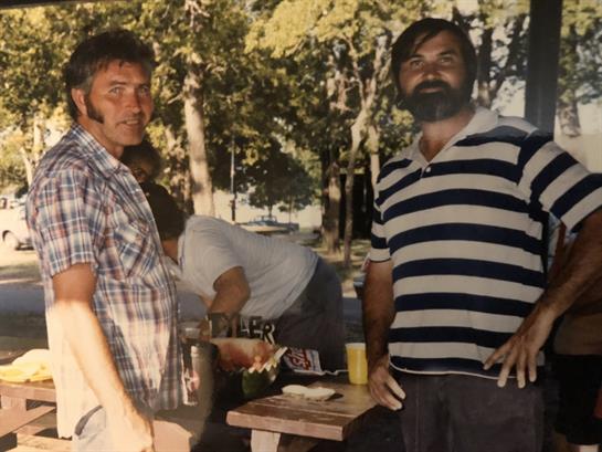 Two men stand at a table in a vibrant outdoor setting, talking and enjoying each other's company.