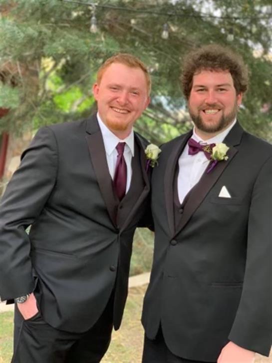 Two smiling men dressed in formal suits stand together outdoors, enjoying a wedding moment.