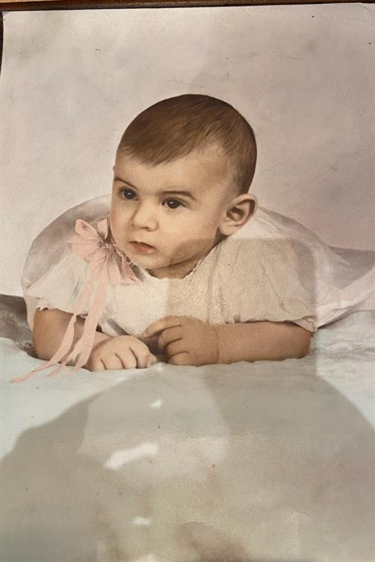 A baby lies on a soft surface, curiously exploring while wearing a delicate dress with a ribbon.