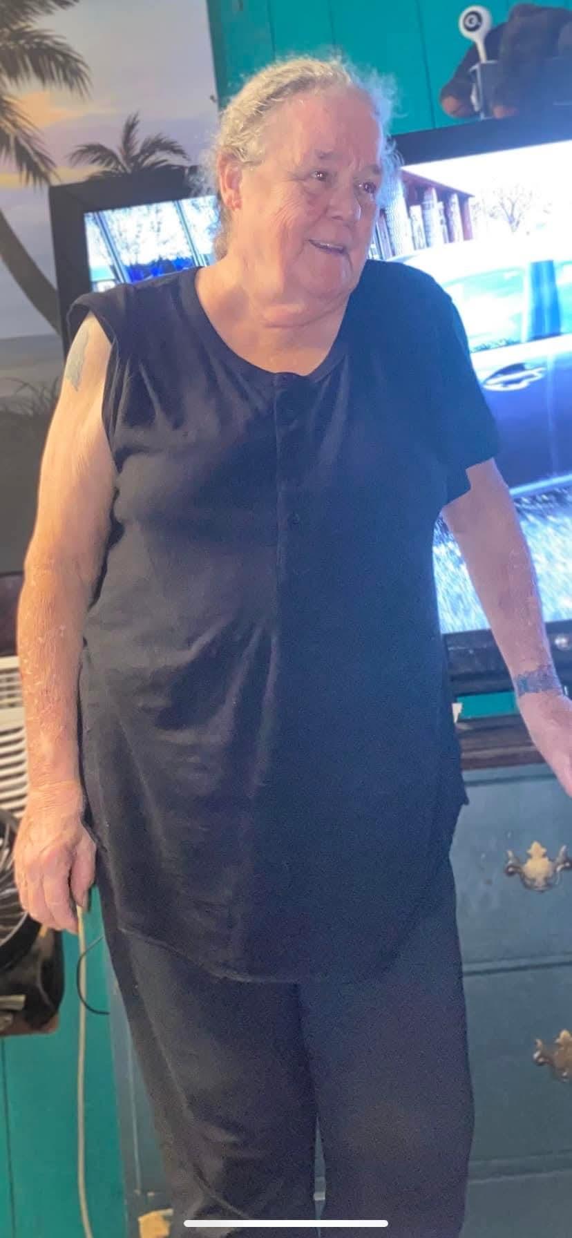An elderly woman stands in her living room wearing a simple black outfit, smiling warmly at guests.