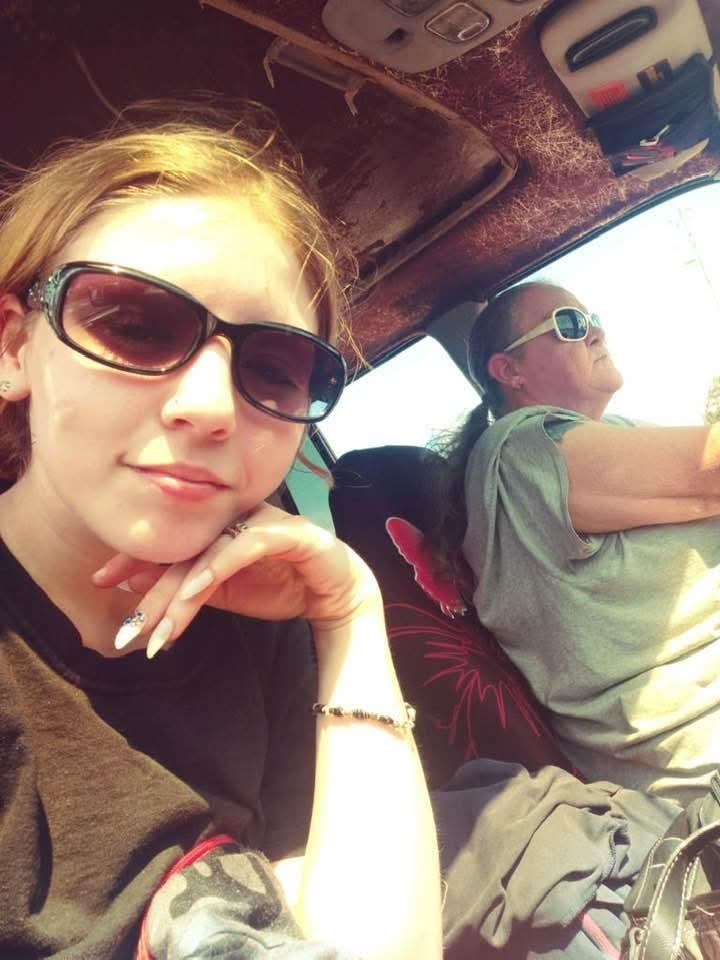 A teenager poses for a selfie while an adult drives, both wearing sunglasses and smiling.