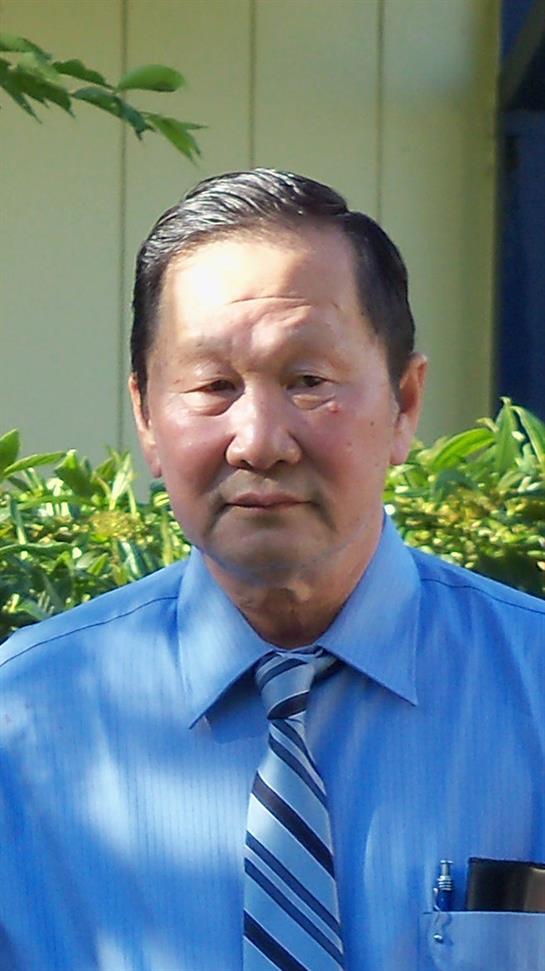 A senior man in a blue shirt and striped tie stands outdoors, surrounded by vibrant green plants.