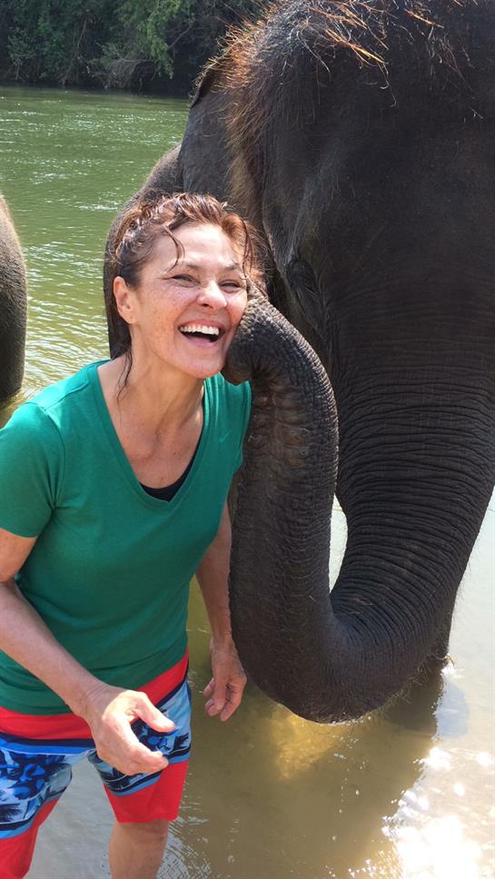 A woman shares a happy moment with an elephant, enjoying nature by the river on a sunny day.