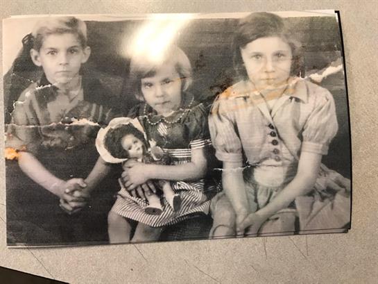 Three children sit together, one holding a doll in mid-1900s attire.