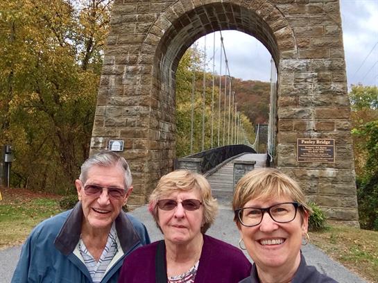 Friends pose happily near a stone bridge framed by fall foliage, enjoying a beautiful day outdoors.