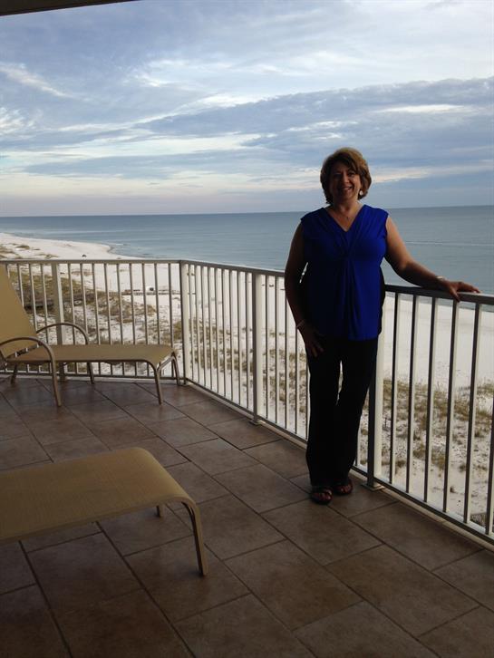 A woman in a blue top stands on a balcony overlooking the tranquil ocean at sunset.