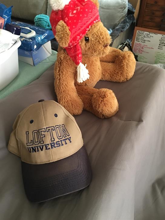 A teddy bear wearing a red hat sits next to a university cap on a gray fabric surface.
