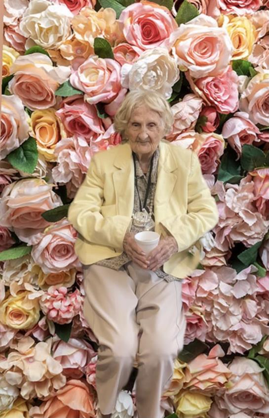 An elderly woman smiles while holding a cup, surrounded by vibrant roses at a floral exhibition.