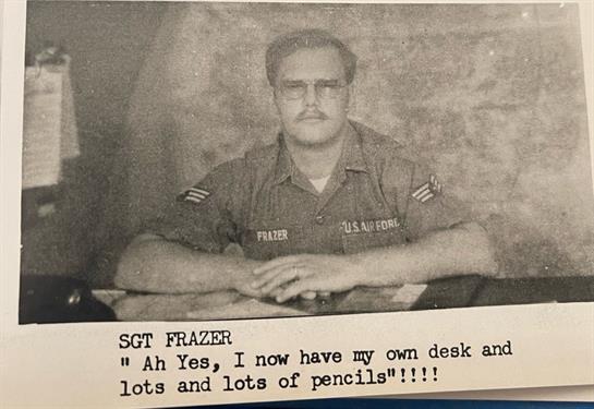 A military veteran sits at a wooden desk, proudly displaying his abundance of pencils while smiling.