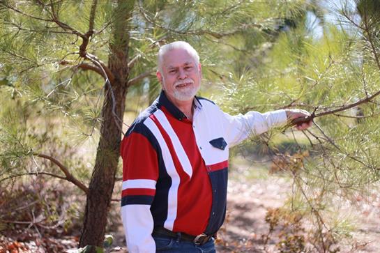 Man enjoys the outdoors, posing happily beside a tree in a sunny environment surrounded by nature.