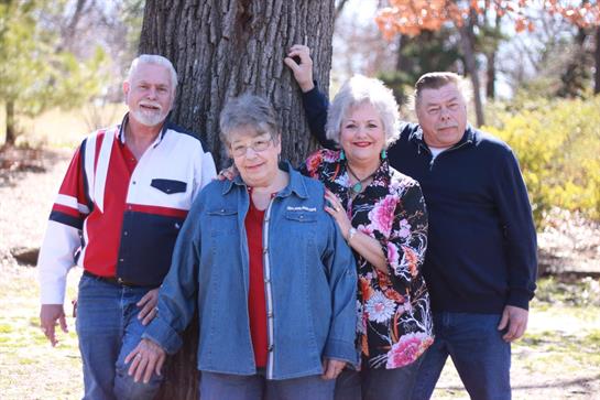 Four adults stand side by side by a large tree in a park, enjoying a sunny autumn day.