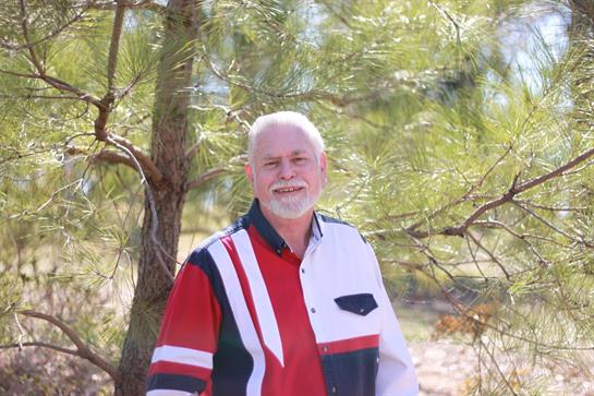 A cheerful man poses in a vibrant shirt, surrounded by pine trees on a sunny day.