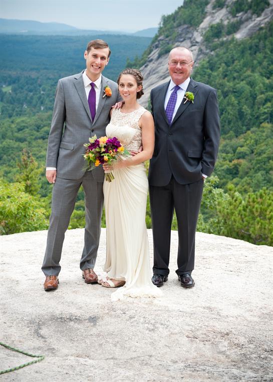 Couple and father happily pose on a rocky ledge with lush mountains behind them at a wedding.