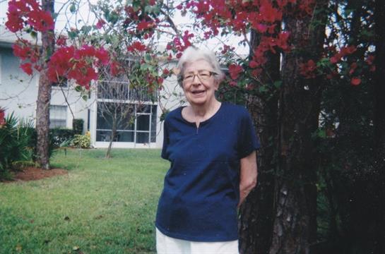 Senior woman stands happily near blooming red flowers in a peaceful garden area.