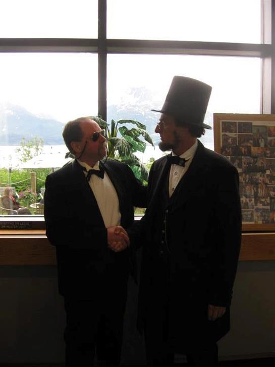 Two individuals in tuxedos interact warmly inside a building with a scenic outdoor backdrop.