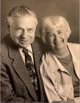 Two elderly individuals pose closely together, both smiling warmly in a studio lit by soft light.