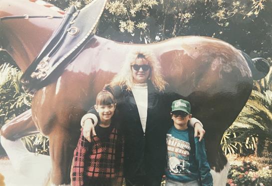 Two children and an adult stand together smiling near a giant horse statue in a park.