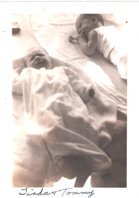 Infant lies peacefully on a blanket with another child visible in the background.