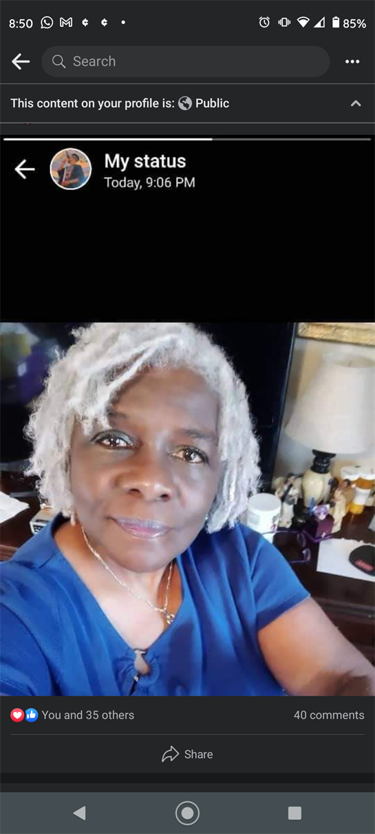 Elderly woman with gray hair smiles warmly while seated in her home, surrounded by personal items.