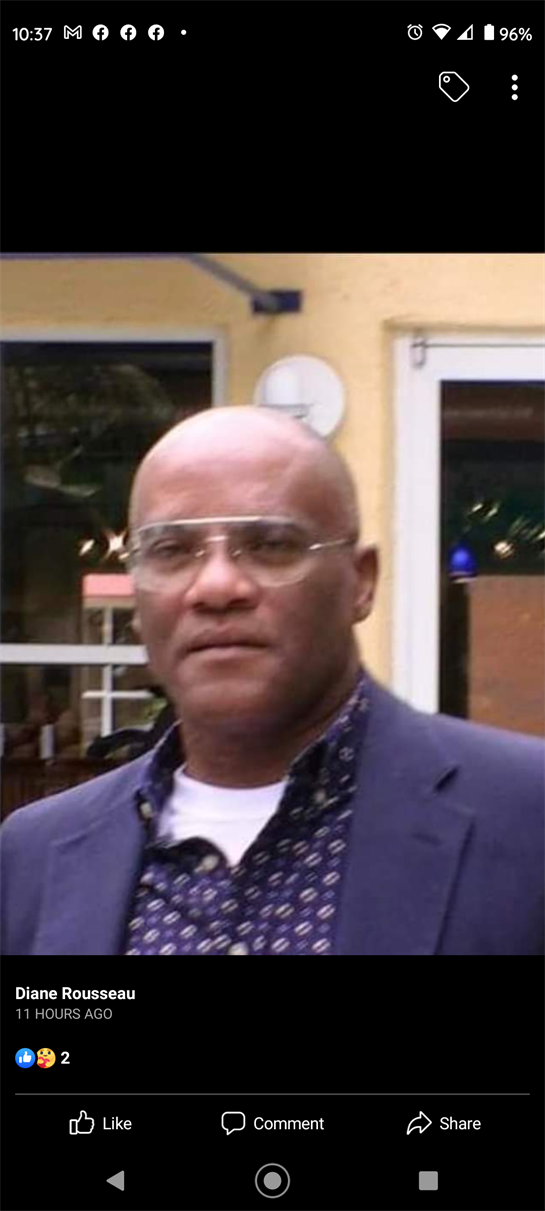 Man in glasses and a blazer stands outdoors, displaying a steady gaze in a lively atmosphere.