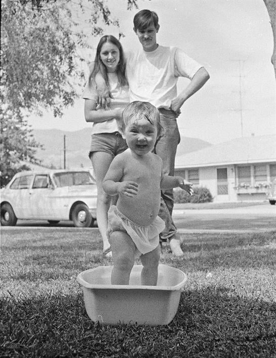 Young child plays in a tub on the lawn as parents observe with smiles on a sunny day.