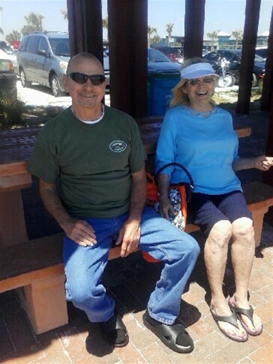 Two seniors sit on a bench at a coastal park, enjoying summer weather and friendship.