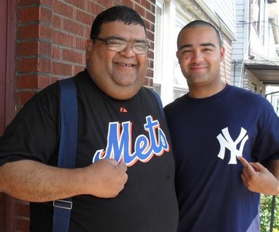 Two friends smile and pose together in baseball jerseys outside a brick building during daylight.