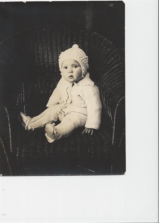 A baby in a cozy knitted outfit sits on a wicker chair, looking curiously at the camera.