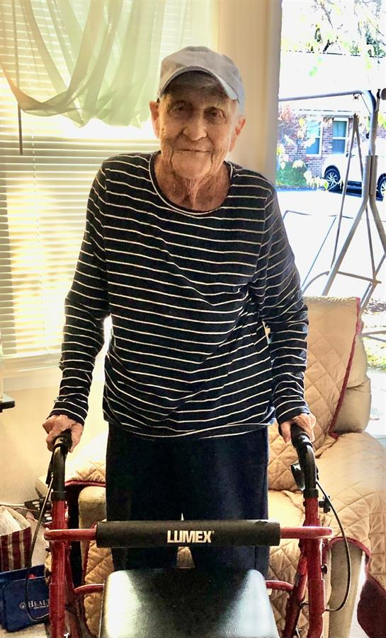 An elderly woman stands with a walker inside a cozy living room, smiling warmly at her surroundings.