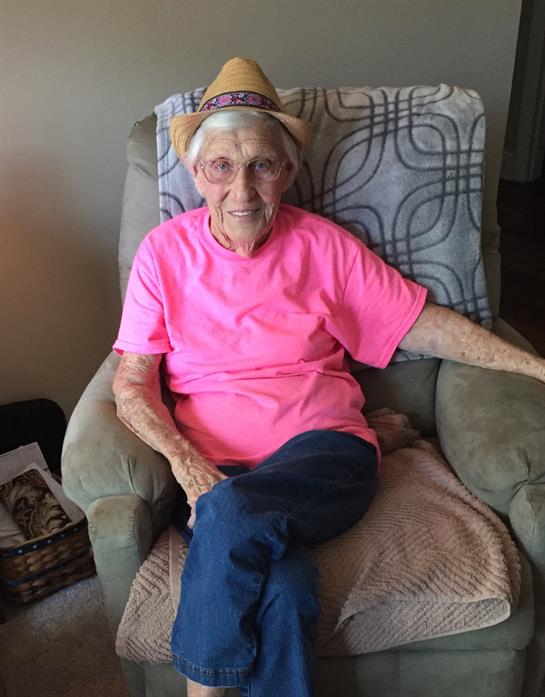 An elderly woman relaxes in a cozy chair, wearing a bright pink shirt and a straw hat.