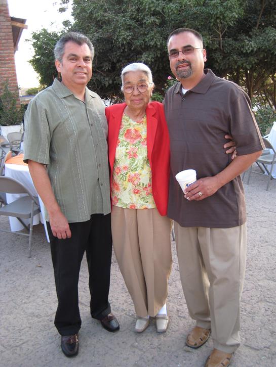 Two men are standing beside an elderly woman in a colorful outfit, enjoying a family celebration.