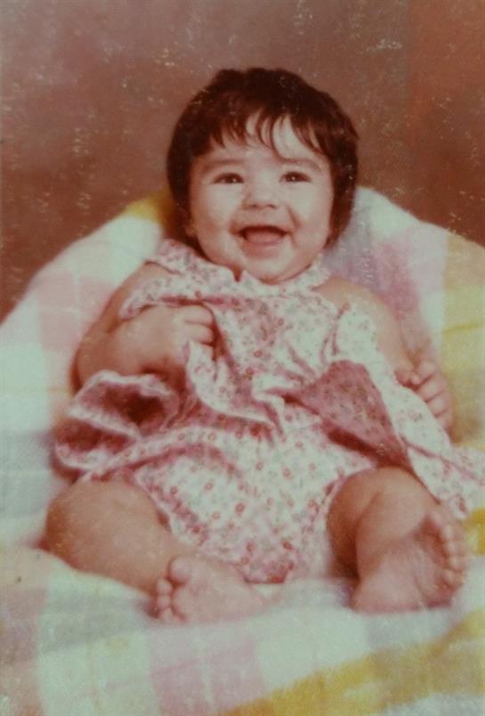 Happy baby wearing a floral outfit holds a small blanket while sitting on a soft surface.