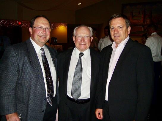 Three men dressed in formal suits smile and pose closely at a social event in the evening.