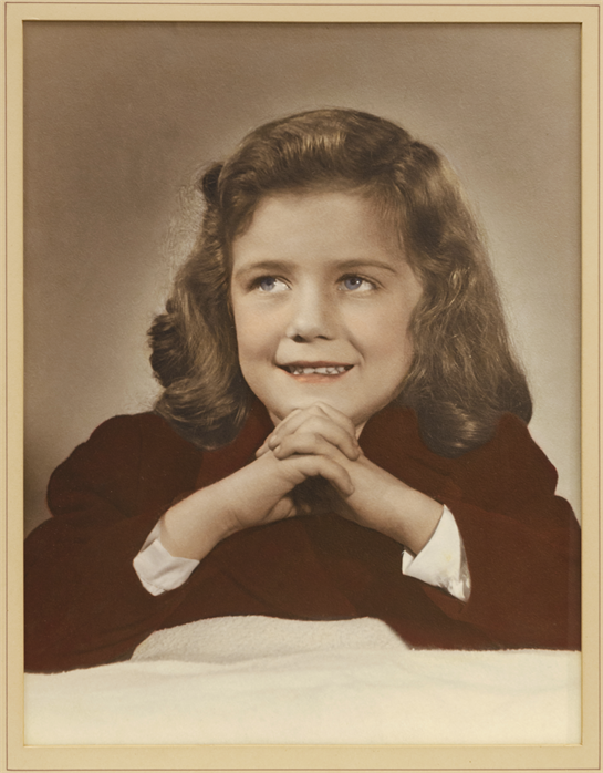 A cheerful young girl sits with her hands clasped, smiling softly at the viewer in vintage style.