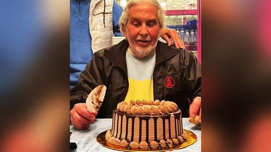 An elderly man happily enjoys his birthday cake, surrounded by friends in a warm setting.