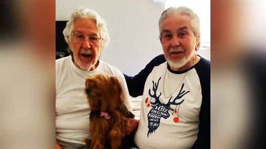 Two older gentlemen sit side by side, smiling and holding a small dog, showing friendship and joy.