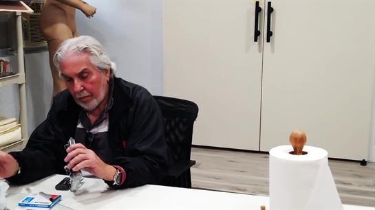 Man with gray hair sits at table, focused on crafting with various tools and materials around him.