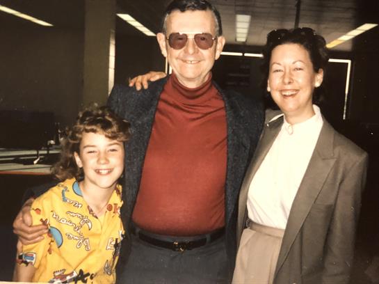 Three people stand together, smiling joyfully in a lively indoor setting during the 1980s.