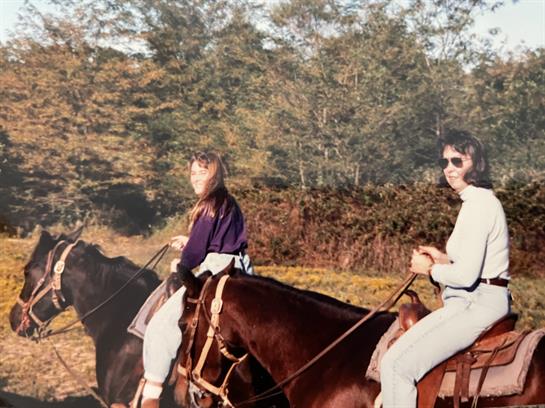 Two women enjoy horseback riding through a vibrant autumn landscape filled with trees and sun.