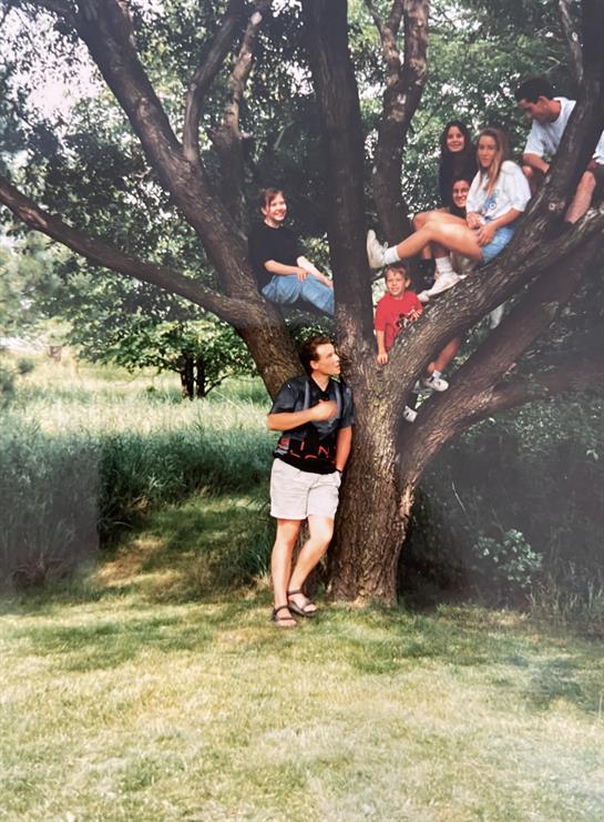 Friends gather in a sunny backyard, some in a tree, others below, smiling and chatting.