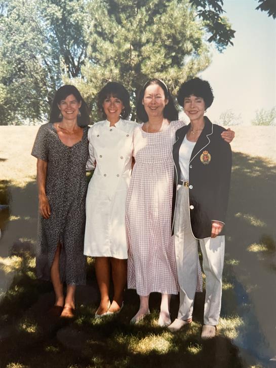 Four women stand together in casual clothing, smiling in a sunny park, enjoying their time together.