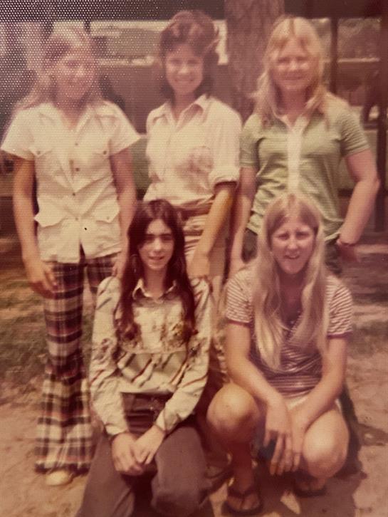 Five young women enjoy a sunny day while posing for a group photo outside in casual attire.