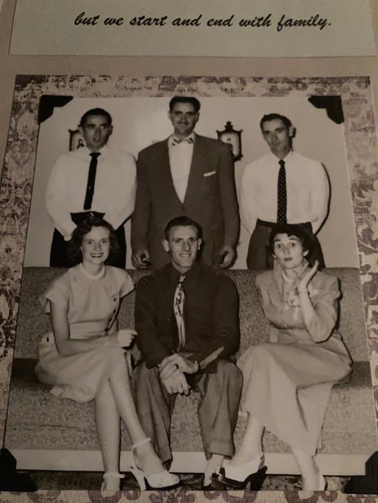 Six individuals dressed in formal attire pose together in a vintage living room setting.