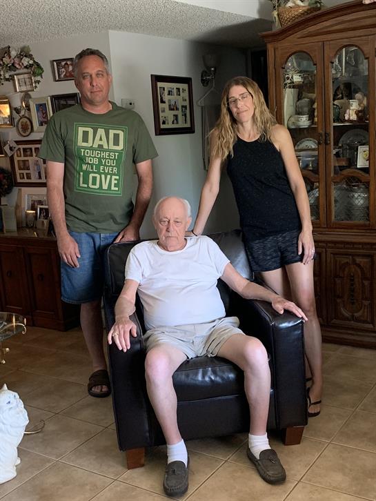 Two adults stand beside an older man seated while capturing a warm family moment in a home.