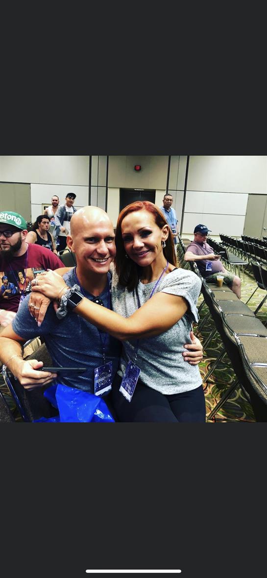 Two people smile and embrace while seated at a conference venue with attendees in the background.