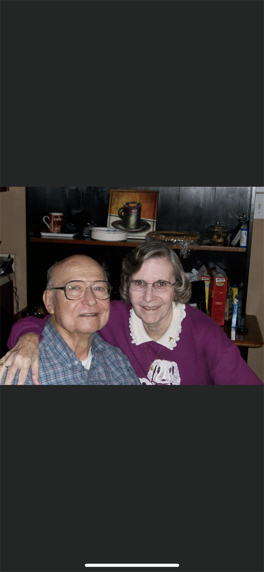 An elderly couple smiles happily together in a warm home environment, surrounded by personal items.