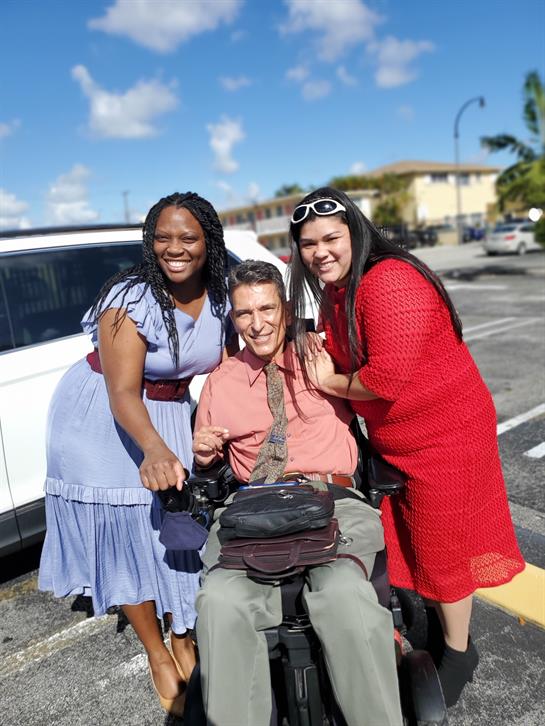 Two women and a man share a joyful moment outdoors, surrounded by blue skies and parked vehicles.