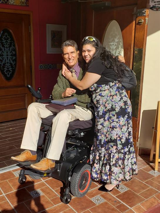 A woman hugs her male friend in a wheelchair outside a welcoming home, radiating happiness.