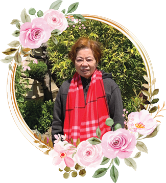 Elderly woman smiles in a garden wearing a red scarf with vibrant flowers in the background.