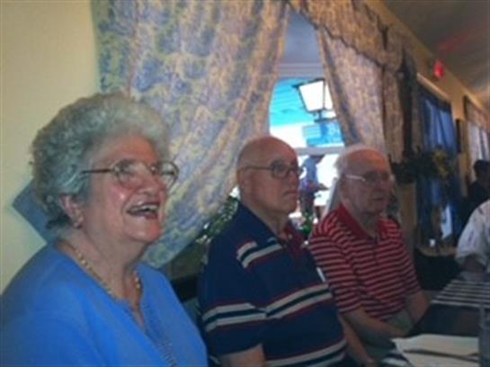 Three elderly individuals are gathered indoors, smiling and engaging in conversation together.
